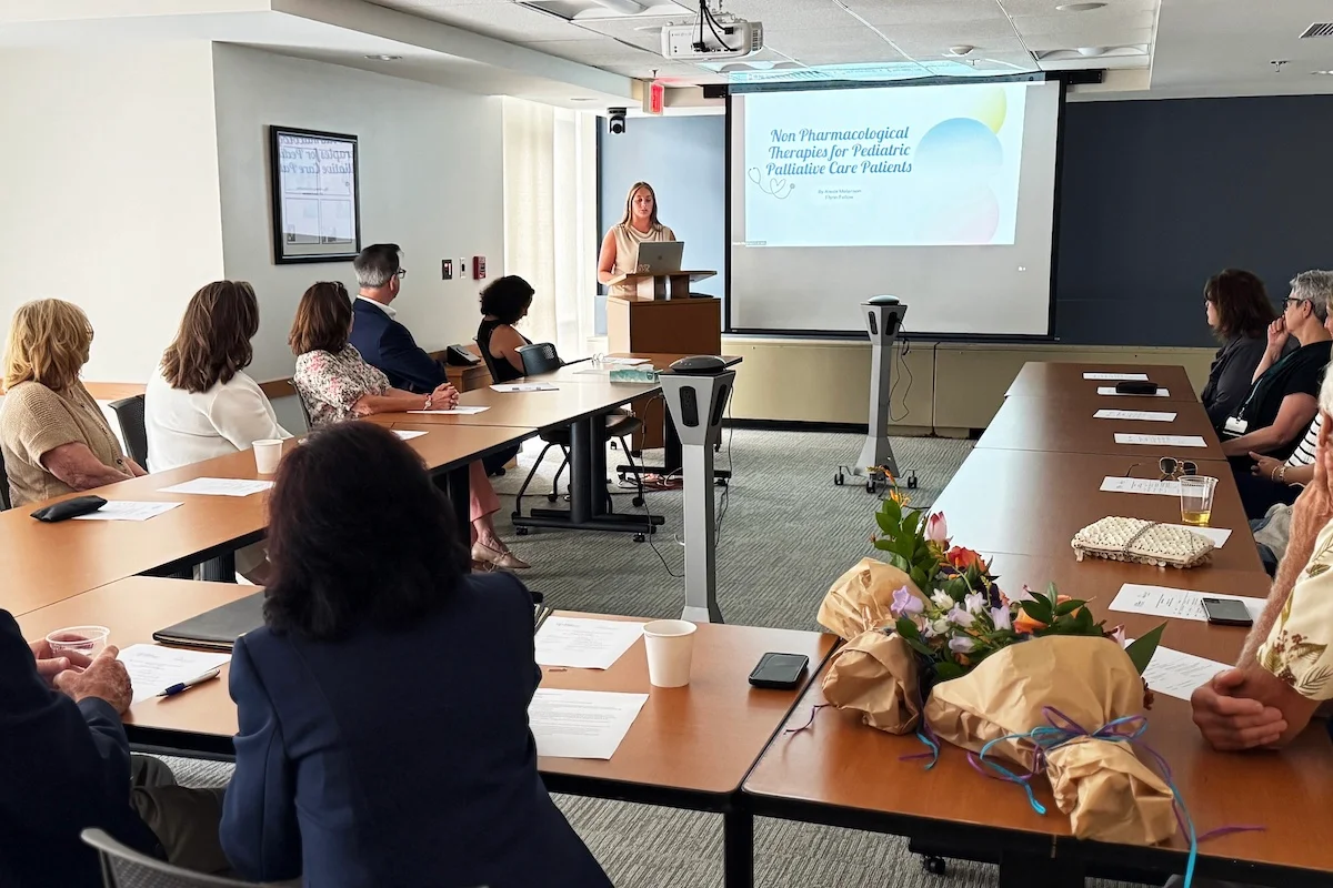 Alexis Melanson standing at a podium in front of a group sitting around a table presenting her research.