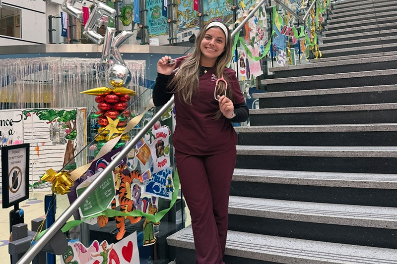 Nursing student Jaidyn Morales-Devine standing on steps smiling with a stethoscope in her hands.