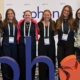 Six professionally dressed women facing the camera smiling.